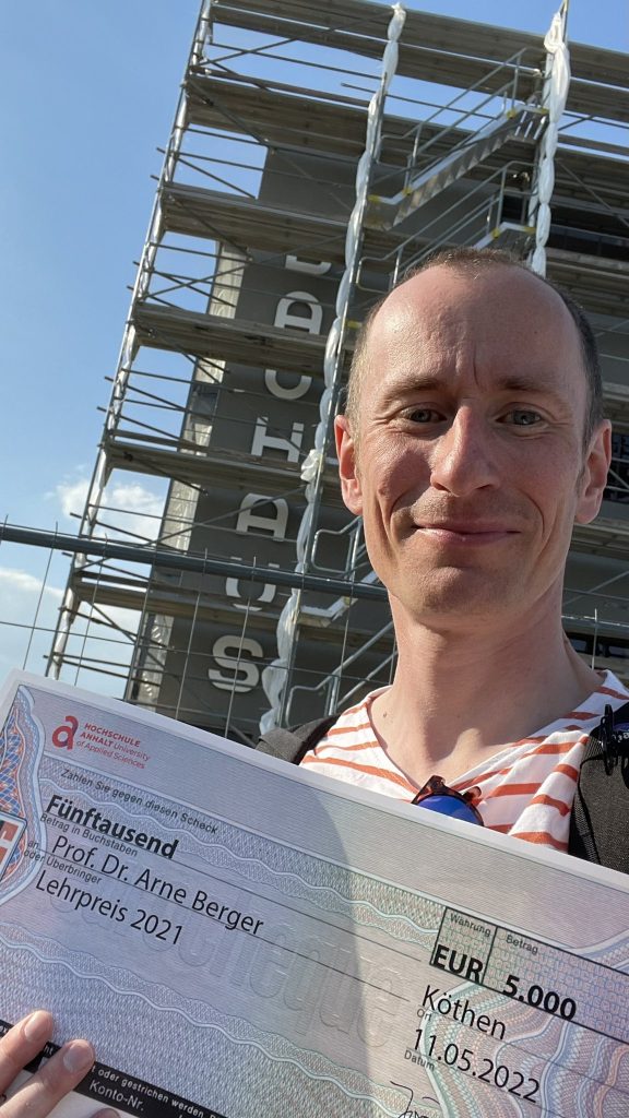 @arneberger attempts to smile and is holding an oversized cheque to the sum of 5000€ in front of the bauhaus in dessau. The sun is shining, the bauhaus is scaffolded.